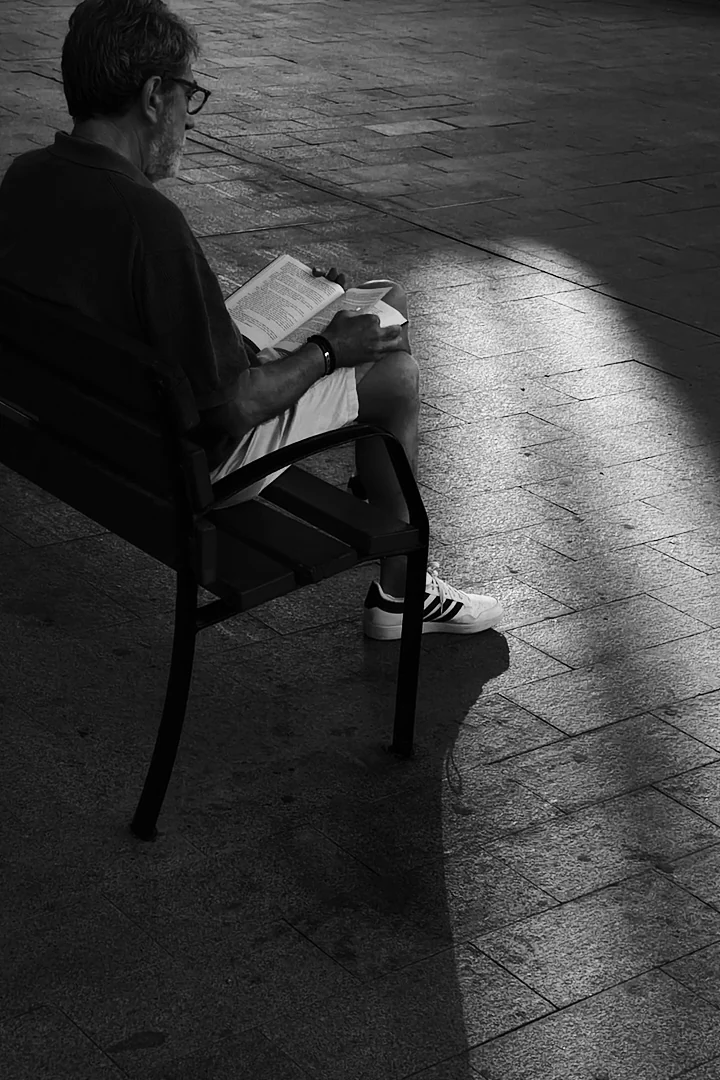 Man seated on a park bench reading a book, a wedge of sunlight falling across the paving beside him