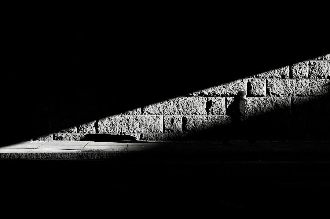 Figure walking along a sunlit stone wall beneath a sharp diagonal of shadow