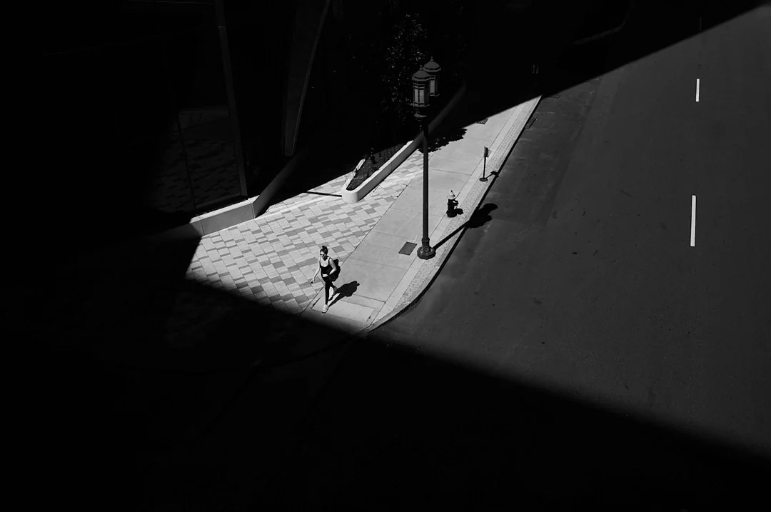 Elevated view of a lone runner crossing a sunlit sidewalk between deep diagonal shadows