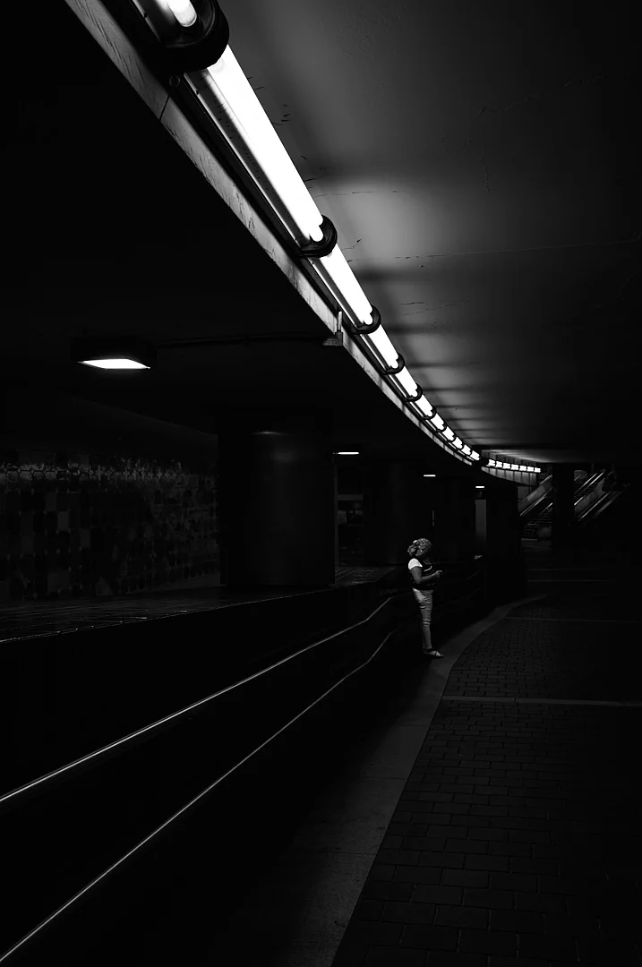 Single figure waiting beside the tracks in a dim station, a curve of fluorescent light overhead