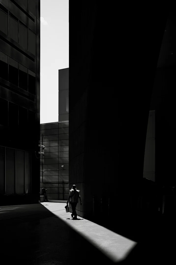 Person with a bag walking through a narrow band of sunlight between tall dark buildings
