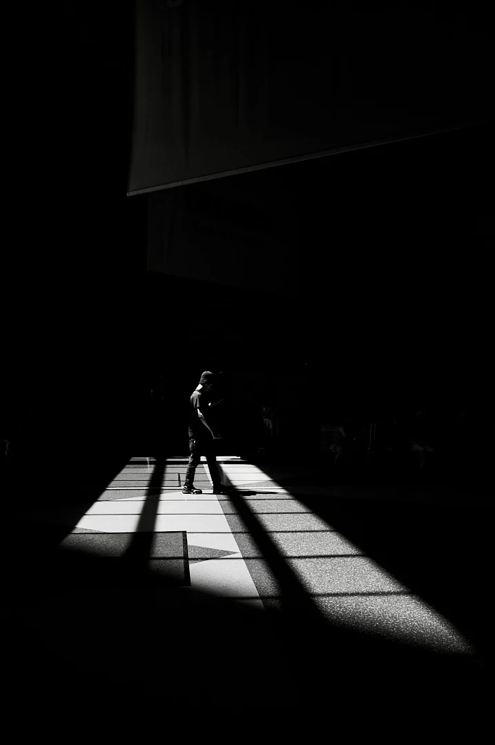 Lone figure crossing a bright patch of pavement, long shadows reaching toward the camera