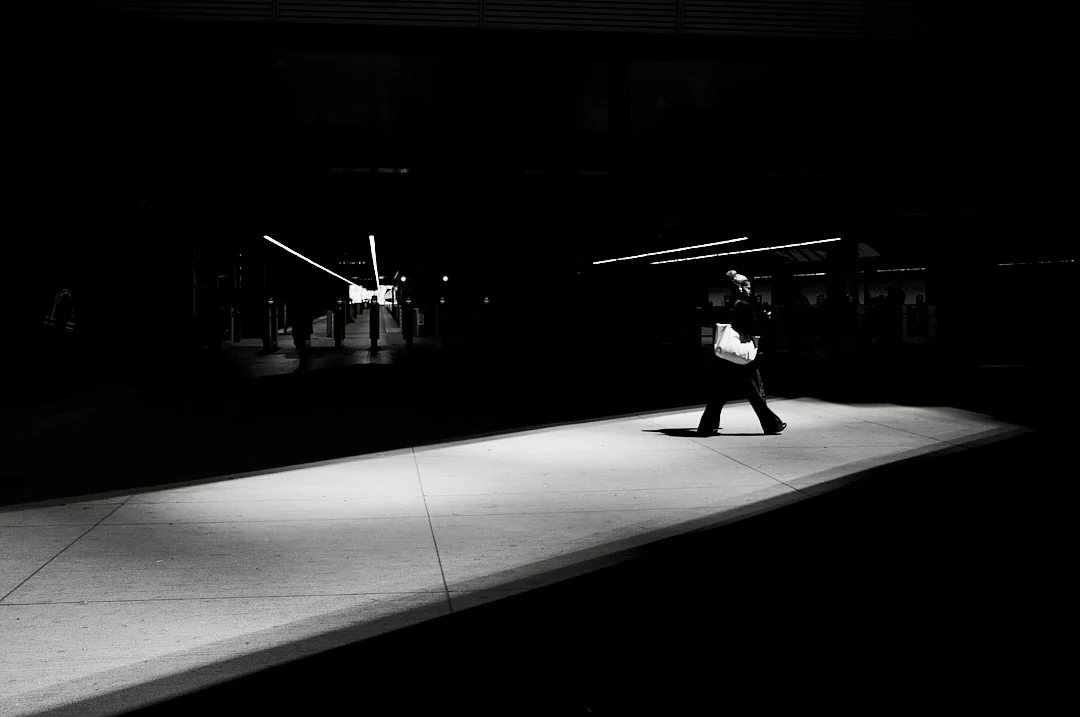 Person carrying a bag walks through a bright strip of sunlight on a dark city sidewalk