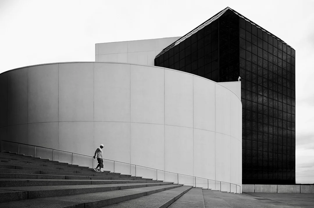 Figure climbing a wide staircase beside a curved white wall and a dark glass tower