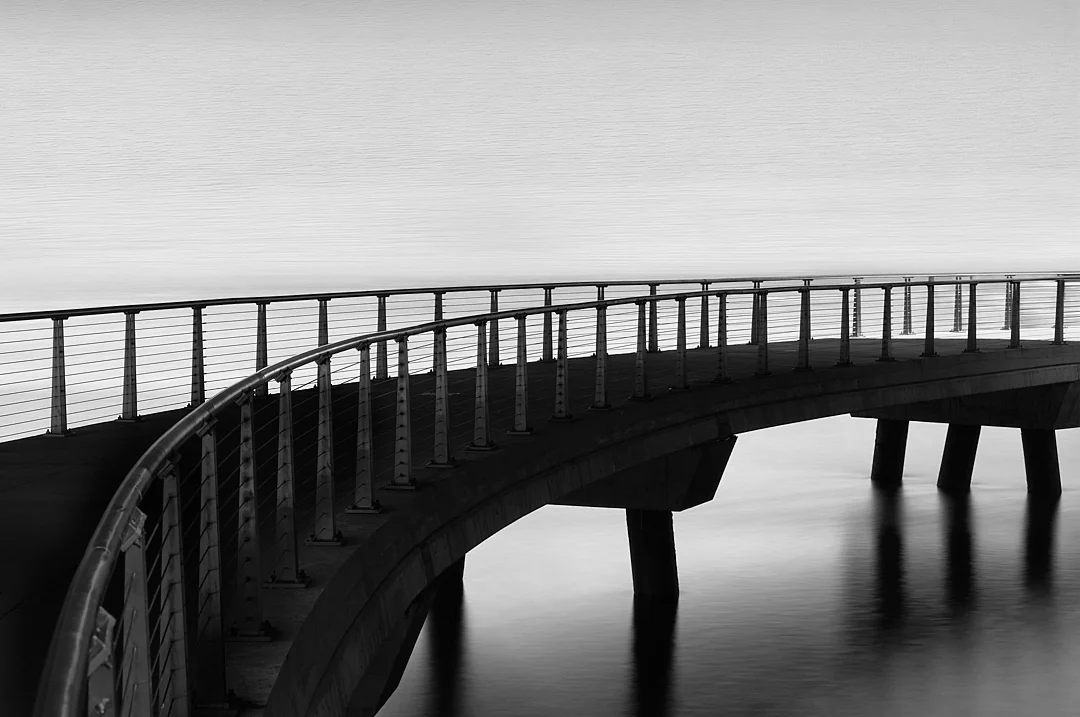 Curving footbridge with railings reaching out over still water under a pale sky