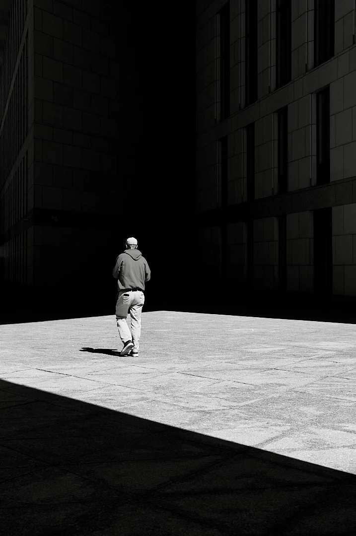 Man in a hooded top walking across a sunlit plaza, buildings dark on both sides