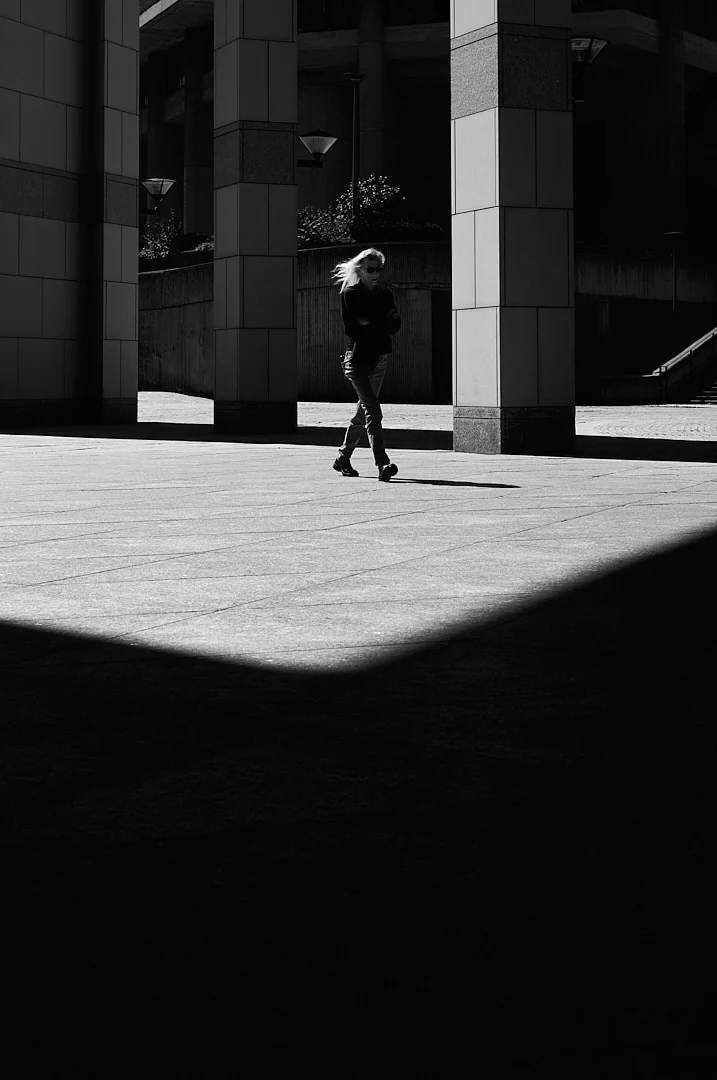 Woman with pale hair walking past stone columns, a sharp shadow cutting the foreground