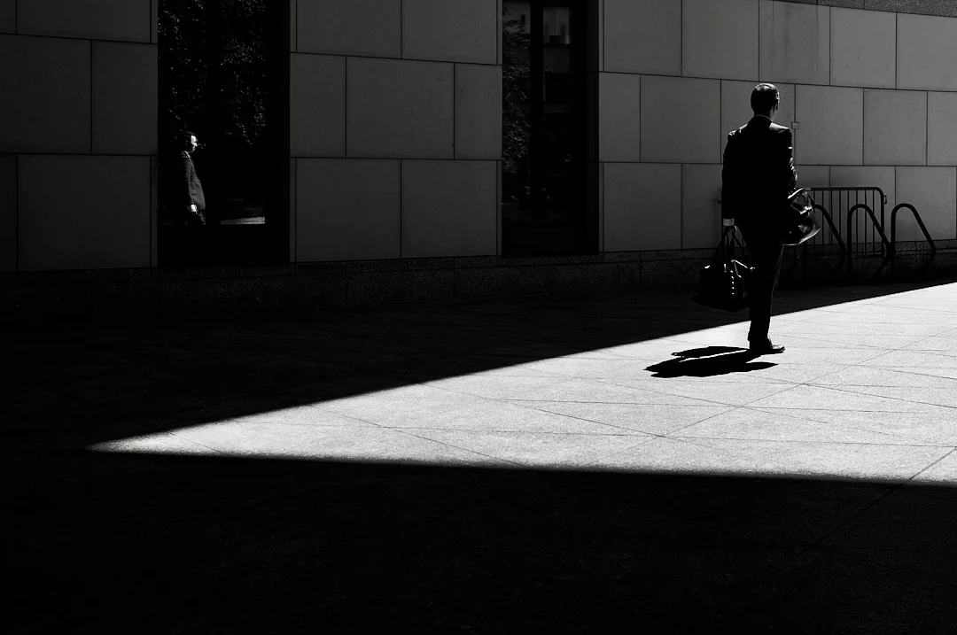 Man with a bag walking along a sunlit wall, his shadow stretching across the plaza