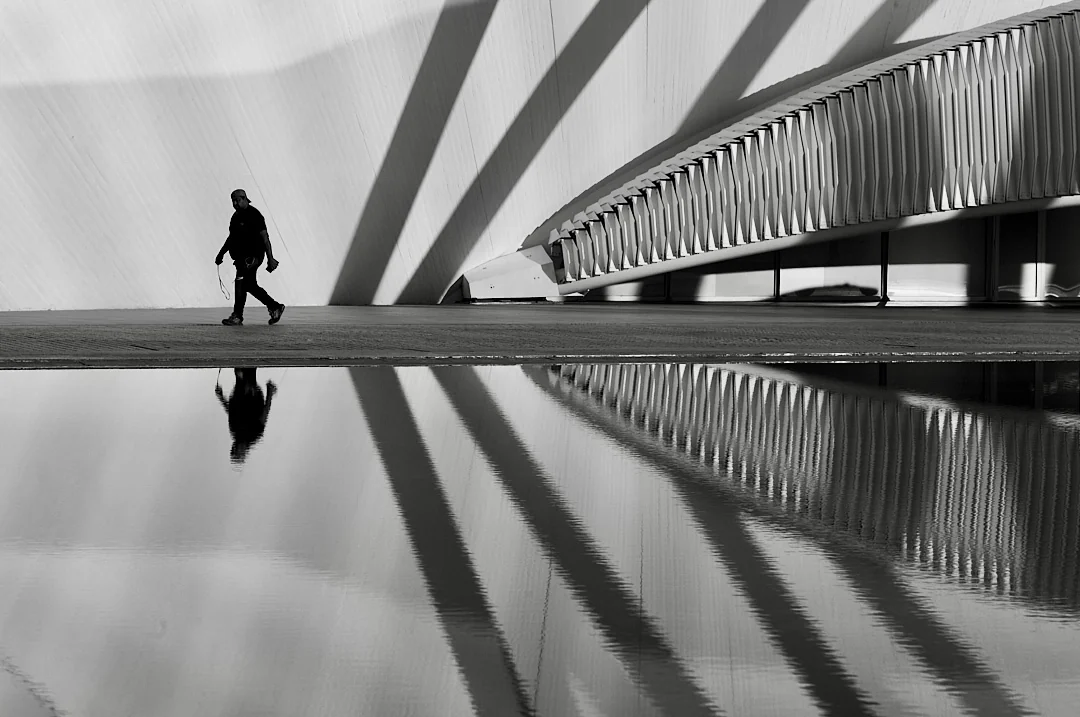 Man walking beside still water, a curved bridge and its reflection behind him