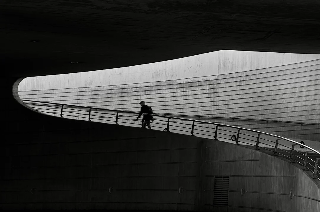 Figure crossing a curved pedestrian bridge beneath a sweeping concrete canopy