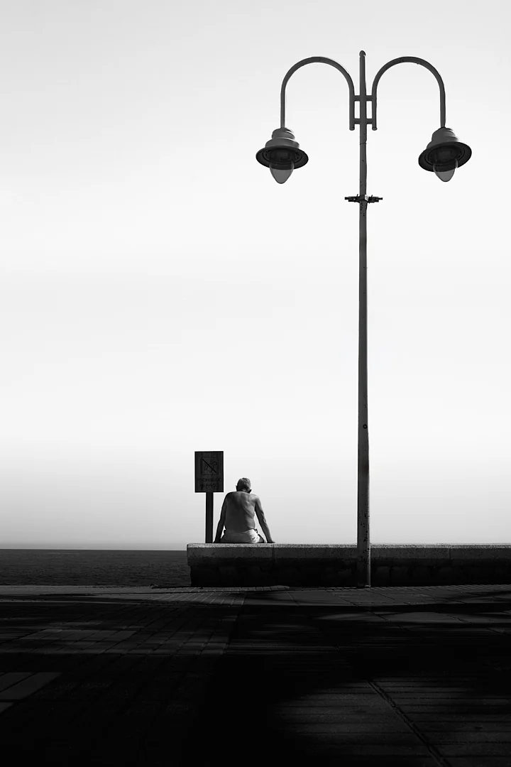 Person sitting alone on a seafront wall beneath a tall double lamp post
