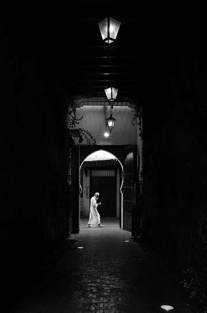 Robed figure passing a lit archway at the end of a dark passage hung with lanterns