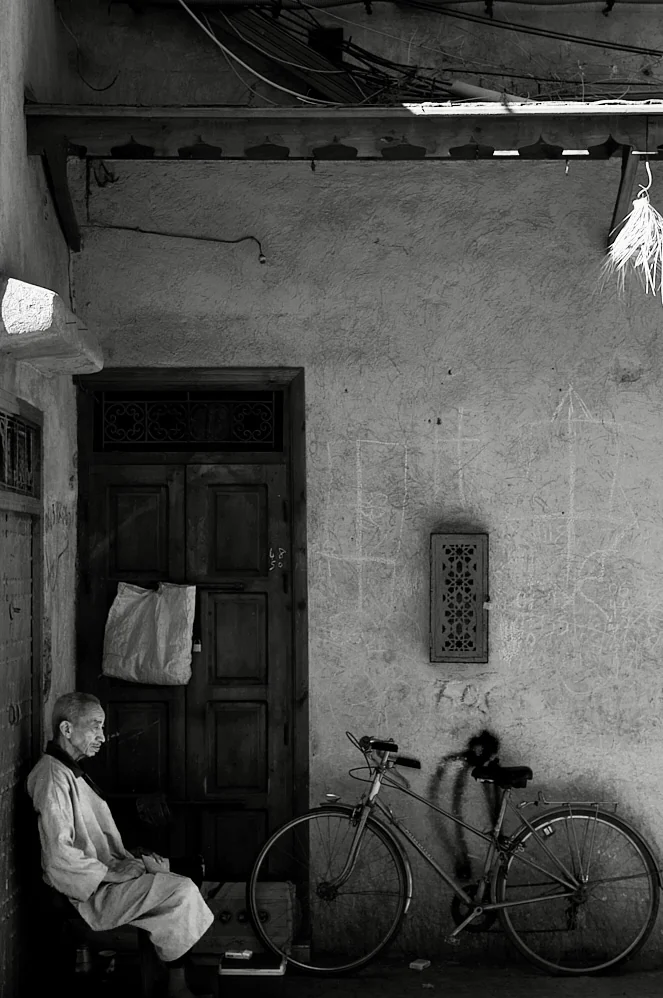 Older man sitting beside a bicycle against a weathered wall with a wooden door