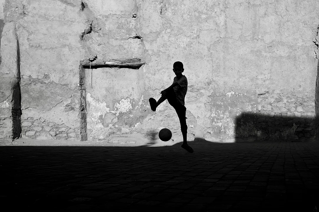 Boy kicking a football against a crumbling wall, his shadow large beside him